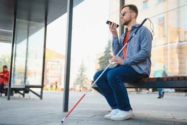 Young blind man with glasses and a cane sitting on bench in park in city, using his smartphone.